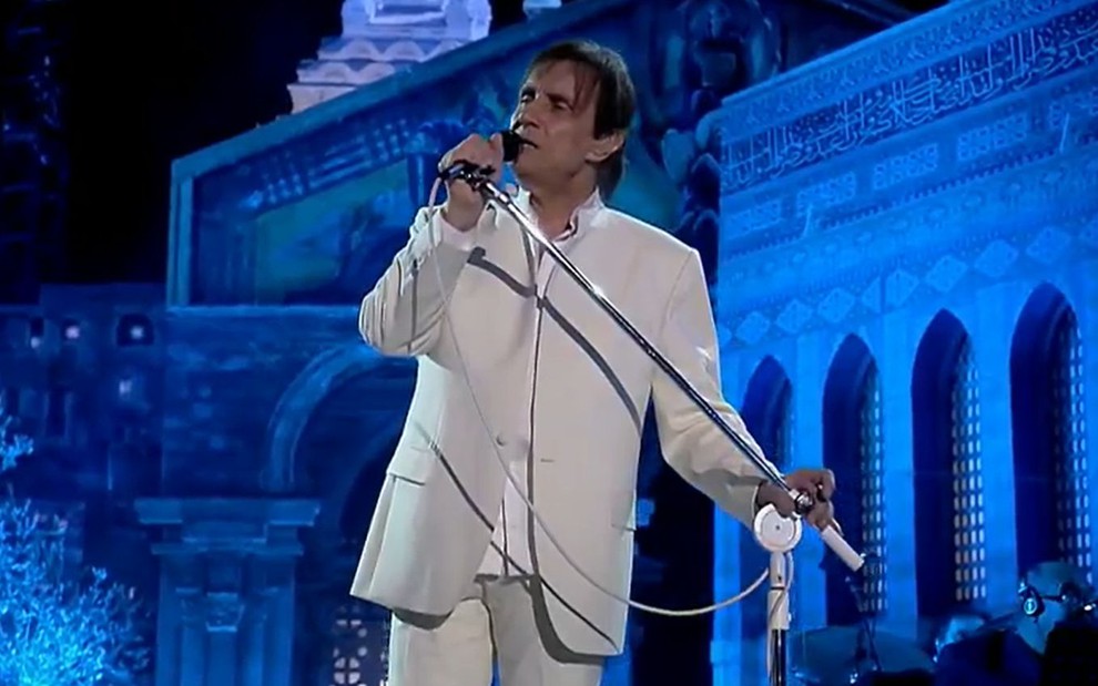 Roberto Carlos de terno na cor gelo segura microfone e canta em cenário de especial em Jerusalém