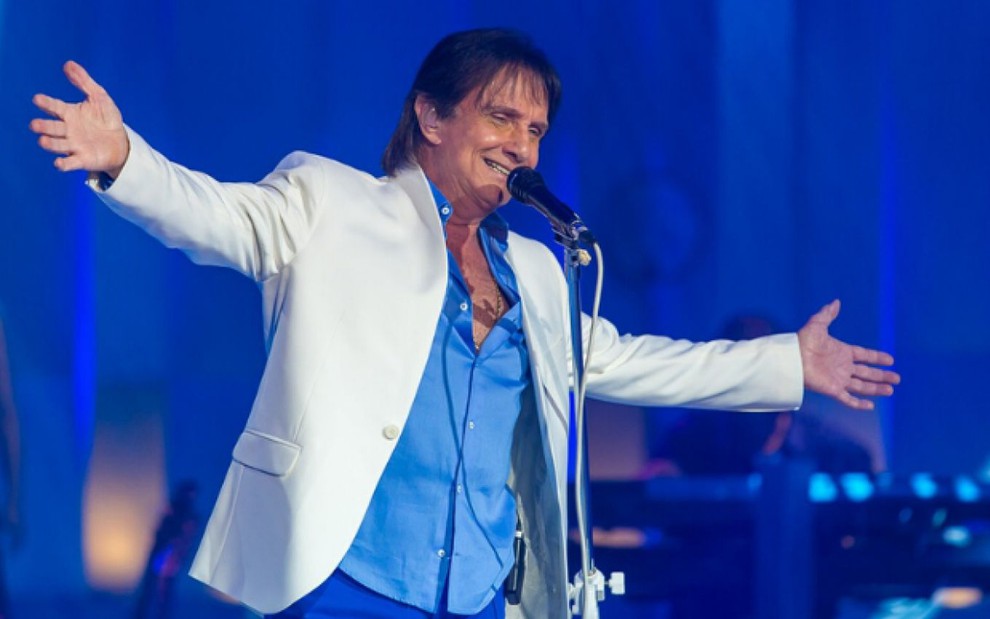 Roberto Carlos de braços abertos usando paletó branco e camisa azul em frente ao microfone durante show