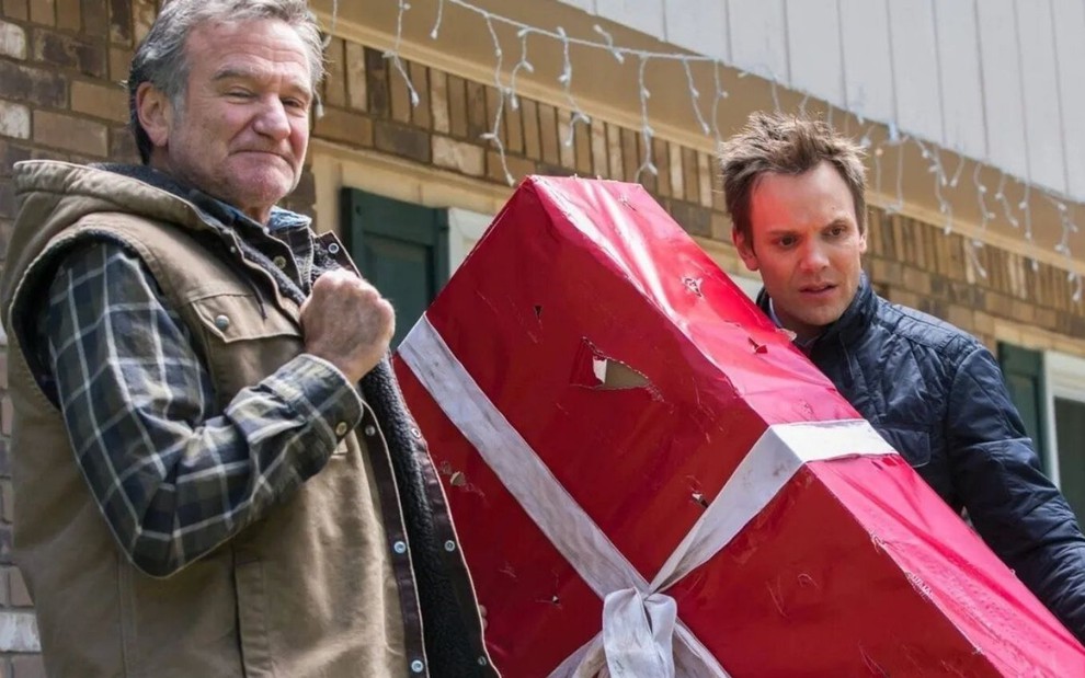 Robin Williams observa Joel McHale carregando um presente em cena do filme Maldito Feliz Natal (2014)