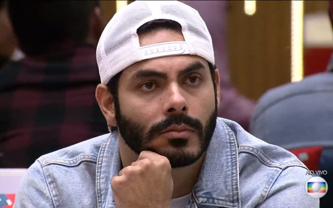 Rodolffo Matthaus de boné branco, jaqueta jeans e mão no queixo, olhando para a câmera
