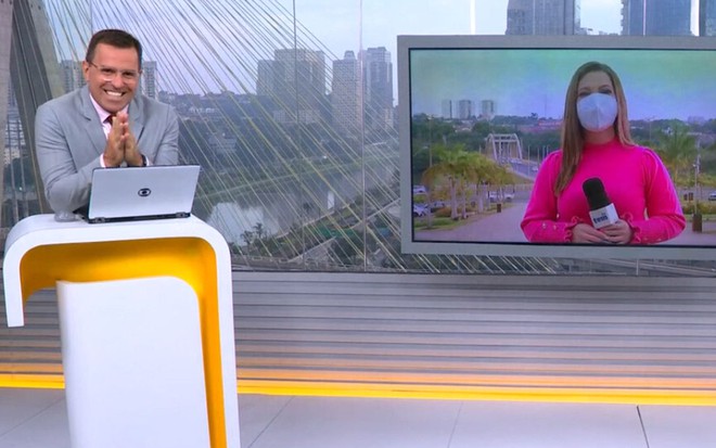 Rodrigo Bocardi em pé na bancada, de terno cinza, sorridente, com as mão juntas; repórter Monize Poiani, de máscara branca e blusa rosa