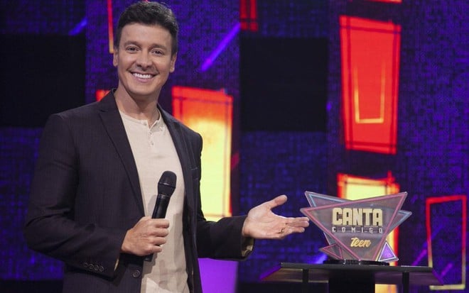 Rodrigo Faro sorridente, de terno preto e camisa branca no palco do Canta Comigo Teen