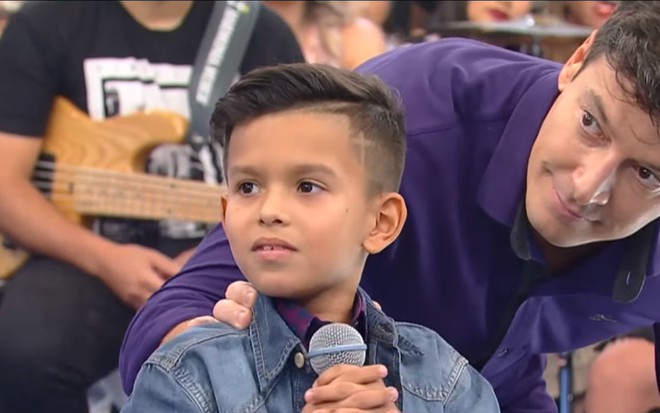 Rodrigo Faro levemente agachado e com uma camisa roxa atrás de um garotinho em seu programa na Record
