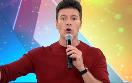 Rodrigo Faro de blusa vermelha, fala ao microfone no cenário de seu programa na Record, Hora do Faro