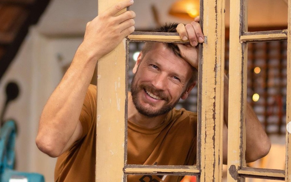 Rodrigo Hilbert sorrindo por trás de uma porta com camisa mostarda, em foto de divulgação do Tempero de Família