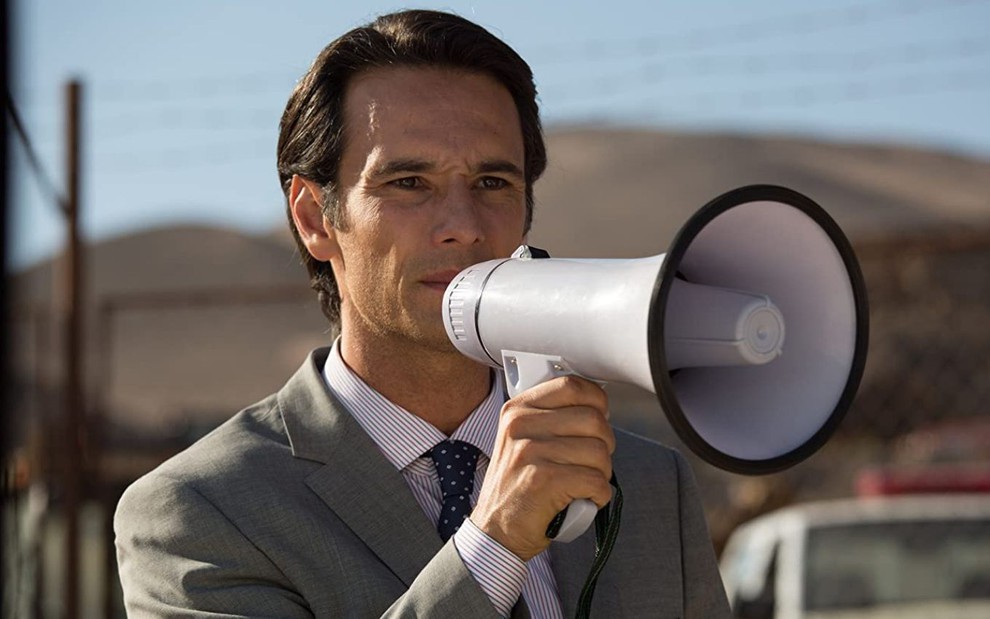 Usando terno cinza, Rodrigo Santoro fala em um megafone em cena do filme Os 33 (2015)