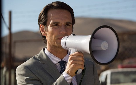 Usando terno cinza, Rodrigo Santoro fala em um megafone em cena do filme Os 33 (2015)