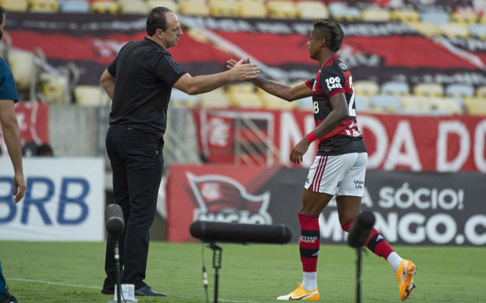 Na lateral do campo Rogério Ceni e Bruno Henrique se cumprimentam após gol do Flamengo, Rogério Ceni está vestido todo de preto e Bruno Henrique vestido com uniforme do time nas cores vermelho, branco e preto