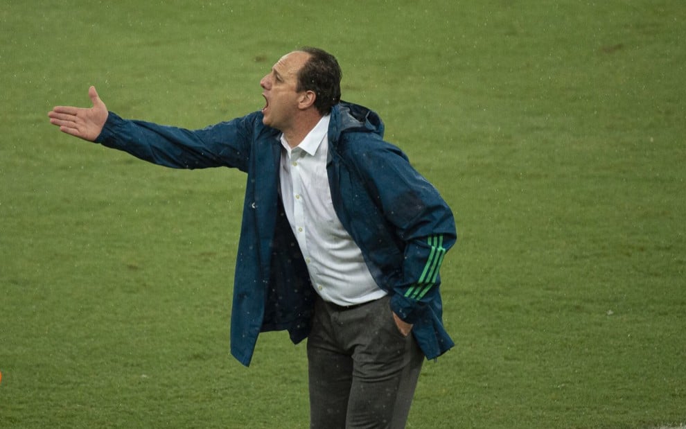 O técnico do Flamengo Rogério Ceni na lateral do campo gritando para comandar o time, ele veste calça e camisa sociais com um agasalho esportivo do Flamengo na cor verde por cima da camisa