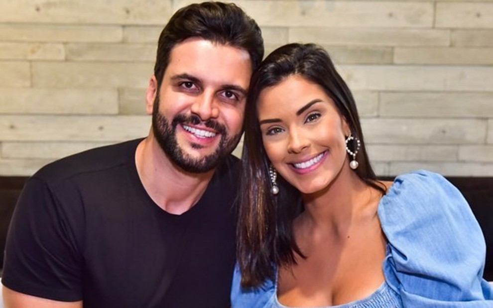 Rogério Fernandes está de camiseta preta; ele aparece ao lado de Ivy Moraes, que veste uma roupa azul; ela usa brincos e está de cabelo solto; ambos sorriem na foto