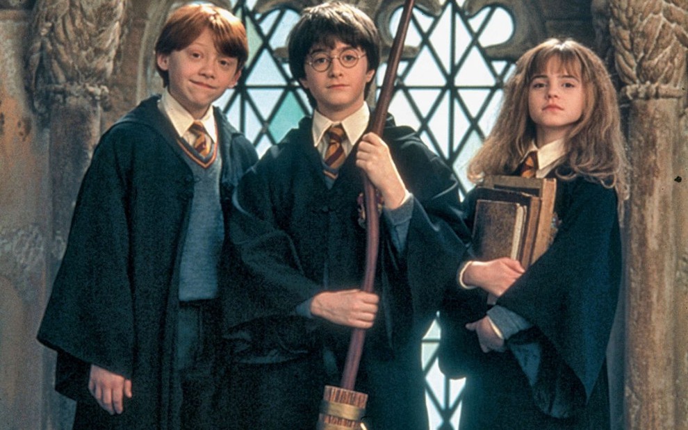 Rupert Grint, Daniel Radcliffe segurando vassoura e Emma Watson com livros antigos na mão