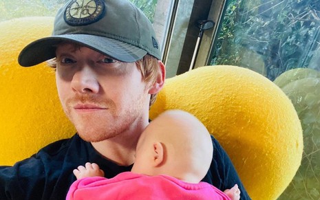 Imagem de Rupert Grint, de boné, segurando a filha no colo