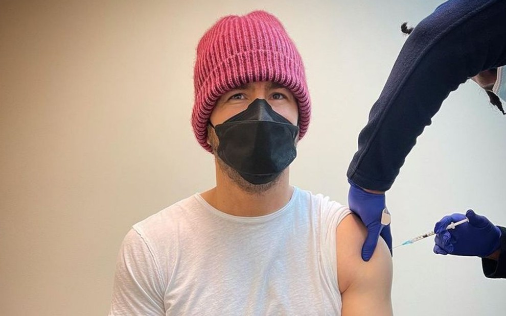 Ryan Reynolds, usando máscara e gorro, recebe sua dose de vacina contra Covid-19 no braço
