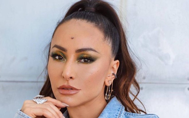 Sabrina Sato está com a mão no queixo, cabelo preso, usando jaqueta azul clara. A apresentadora está encostada em uma parede clara