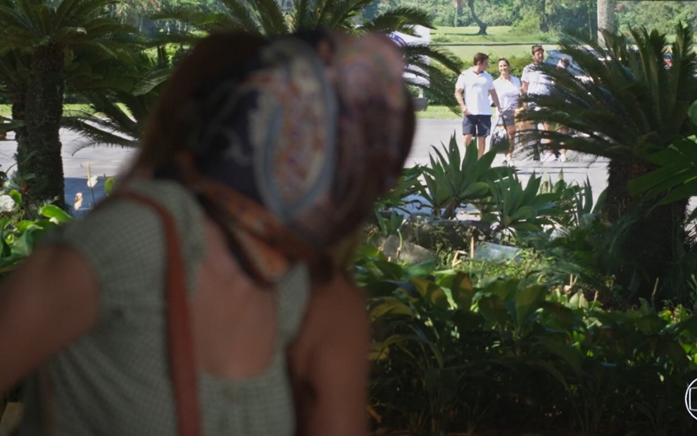 Disfarçada, Deborah Secco observa Rafael Cardoso por trás de um arbusto em Salve-se Quem Puder