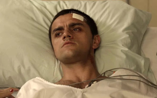 O ator Felipe Simas com expressão bastante séria, em cama de hospital com curativo na testa e fios ao redor, em cena de Salve-se Quem Puder