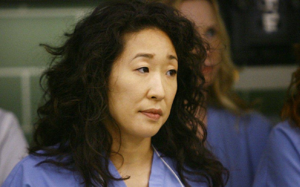 Cristina Yang (Sandra Oh) com avental médico azul e cabelos soltos