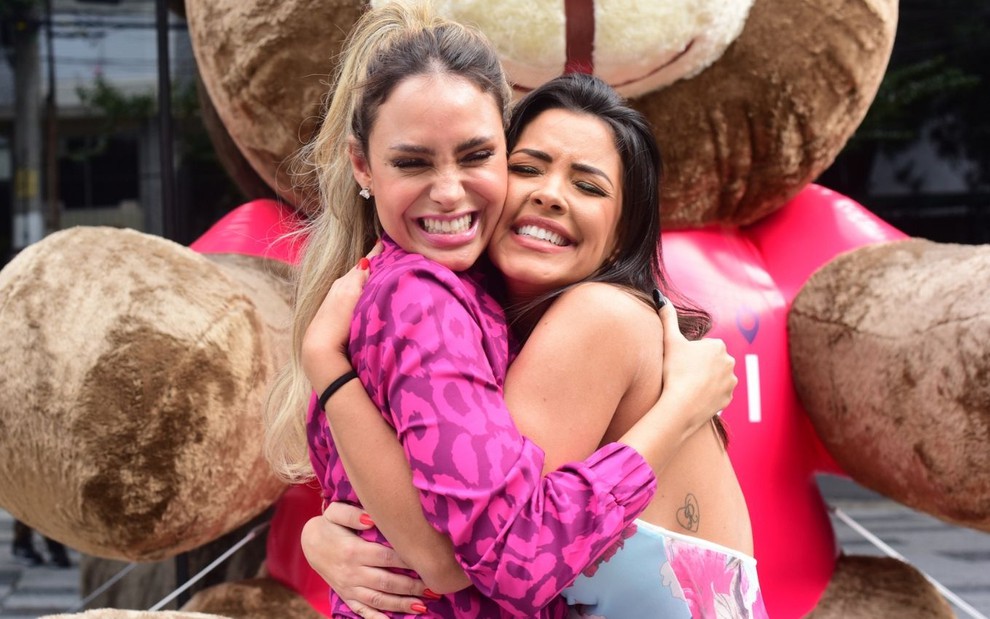Sarah Andrade e Ivy Moraes se abraçam com o rosto próximo em calçada em frente a uma estátua de urso de pelúcia gigante, sem máscara