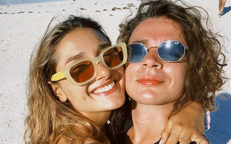 Sasha sorridente abraçada a João Figueiredo na praia; ambos de óculos escuros se protegendo do sol
