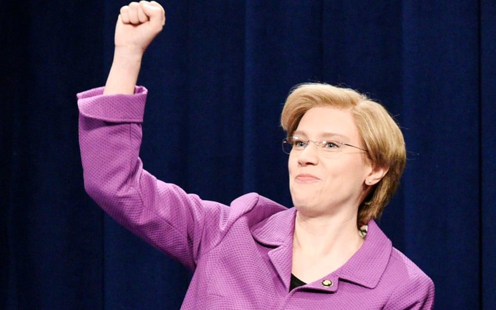 Vestindo um blazer rosa, a atriz Kate McKinnon ergue o braço direito e cerra o punho ao imitar Elizabeth Warren em SNL
