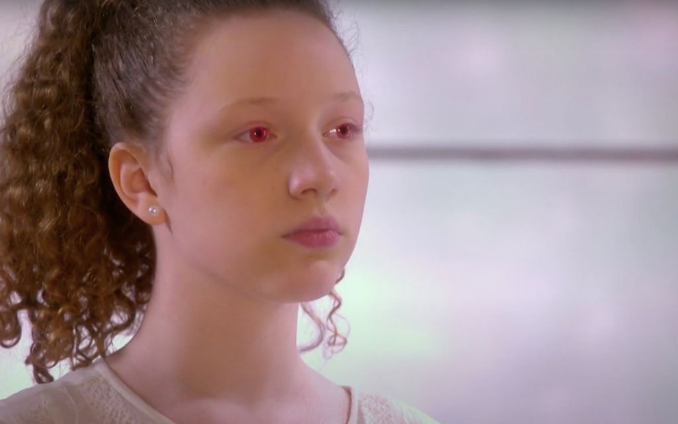 Manuela Kfouri em cena como a personagem Ester, que está com os olhos robóticos vermelhos, em As Aventuras de Poliana