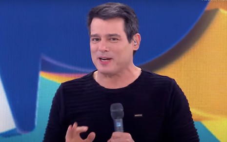 O apresentador Celso Portiolli e blusa preta segura um microfone com a mão esquerda no cenário do Passa ou Repassa