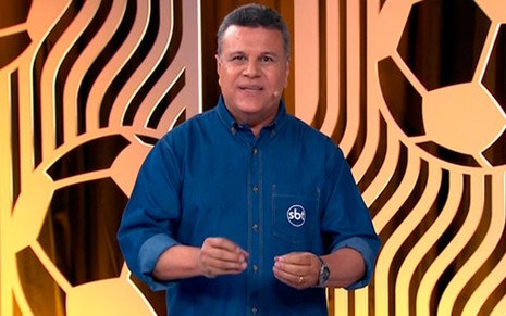 Em um cenário com tons de amarelo e laranja, o narrador Téo José veste camisa azul do SBT na transmissão do Campeonato Carioca