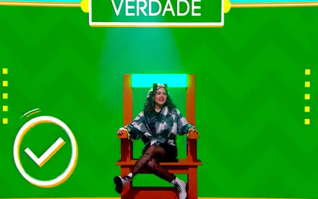 A apresentadora Maisa Silva sentada em uma cadeira elétrica com um fundo verde escrito verdade ao fundo