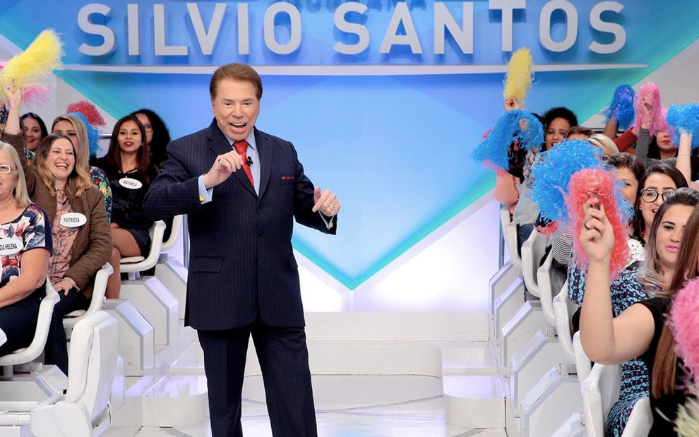 Silvio Santos está com as mãos erguidas a altura do peito com um terno azul e gravata vermelha no meio da plateia do Programa Silvio Santos