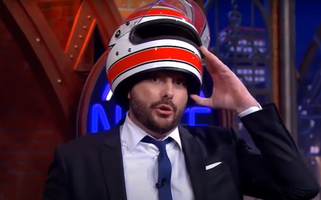 O apresentador Danilo Gentili segura um capacete em cima de sua cabeça durante o programa The Noite, do SBT, exibido na noite de segunda-feira (26)