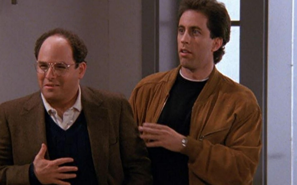 Com a mão no lado esquerdo do peito, Jason Alexander fica surpreso, à frente de Jerry Seinfeld, na comédia Seinfeld