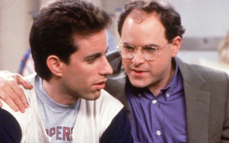 De blazer escuro, o calvo Jason Alexander abraça pelo ombro o colega Jerry Seinfeld em cena da série Seinfeld