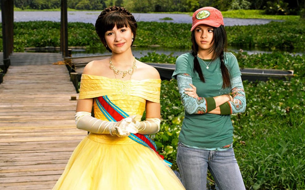Demi Lovato como Rosalinda e Selena Gomez como Carter em Programa de Proteção de Princesas, Demi Lovato está usando uma vestido de princesa amarelo e Selena Gomez está vestindo camiseta verde, calça jeans e boné