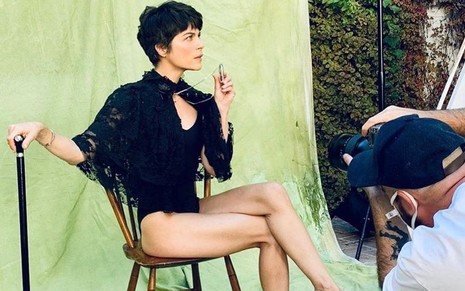 Selma Blair produzida com roupa sensual preta, sentada em cadeira e apoiada a uma bengala no quintal de sua casa