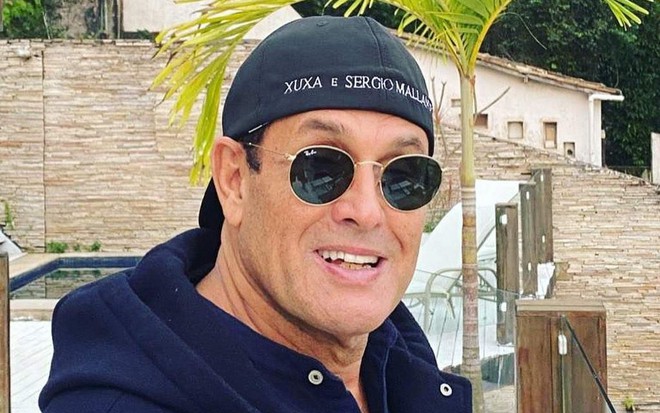Sérgio Mallandro com óculos escuros característicos de seu estilo, casaco azul e boné preto virado para trás