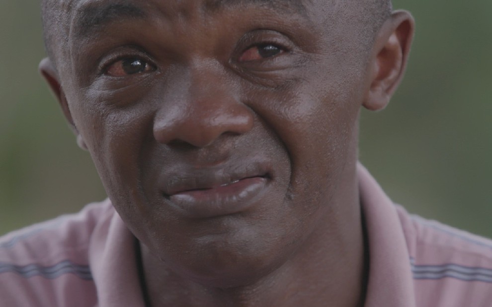 Homem negro com camisa rosa e detalhes em azul tem semblante emocionado e com olhos vermelhos durante relato