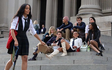 Elenco da nova versão de Gossip Girl sentado em escada observando a chegada de uma personagem em pé no lado esquerdo