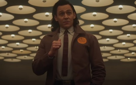 Loki (Tom Hiddleston) em cena da série do Disney+ de camisa branca, gravata e jaqueta apontando para si mesmo com o polegar direito