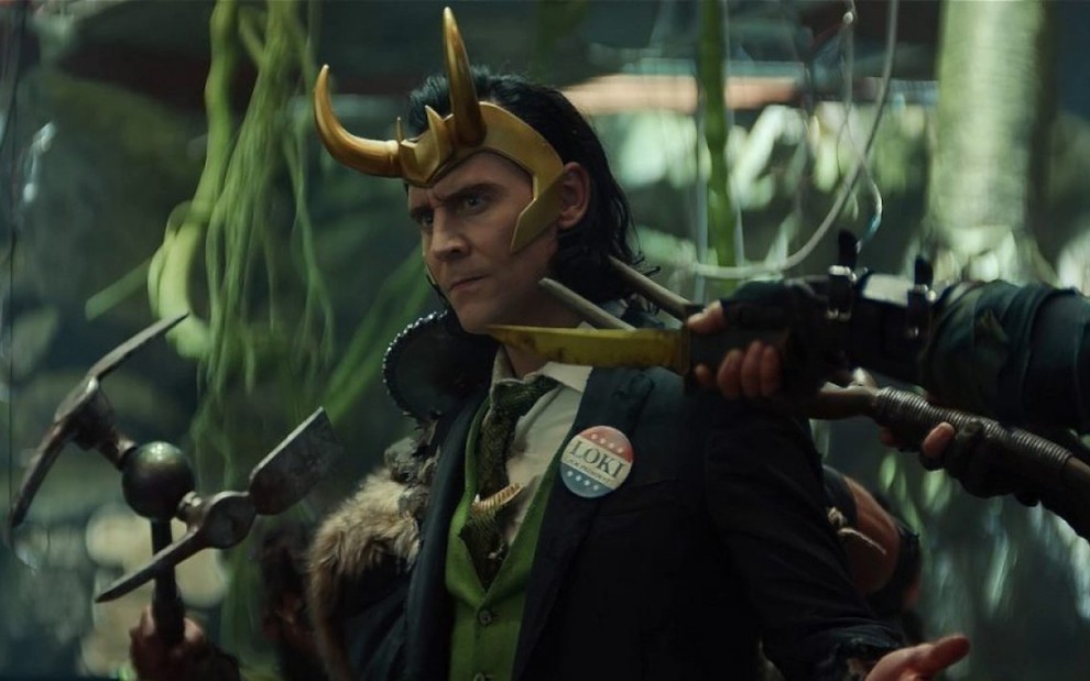 Tom Hiddleston caraterizado como Loki e vestindo um paletó azul sendo ameaçado com uma espécie de faca em sua direção