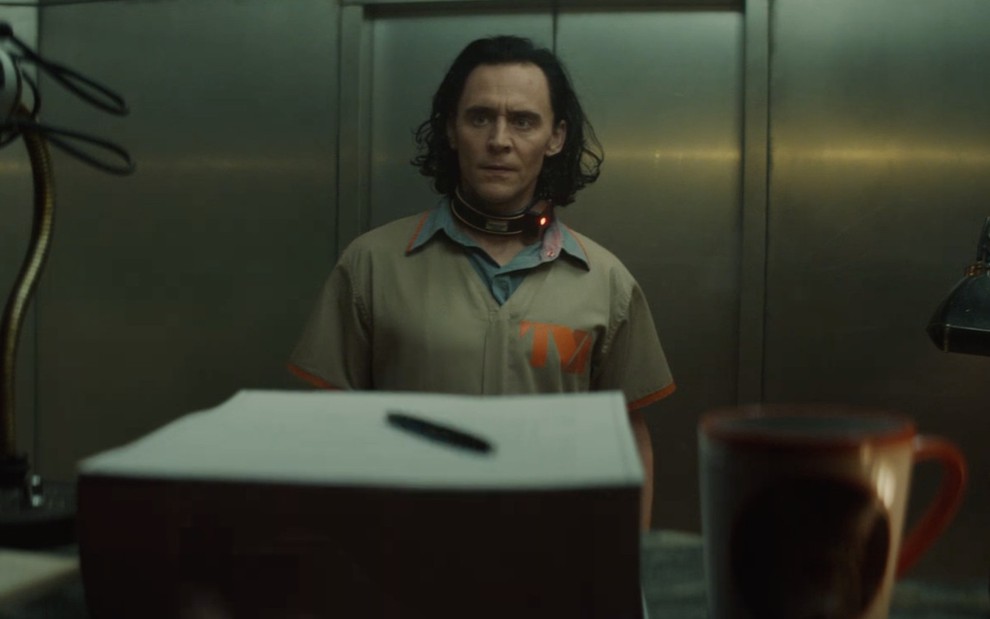 Loki (Tom Hiddlesto) com macacão bege e colar especial no pescoço diante de pilha de papel e uma xícara com fundo de um elevador