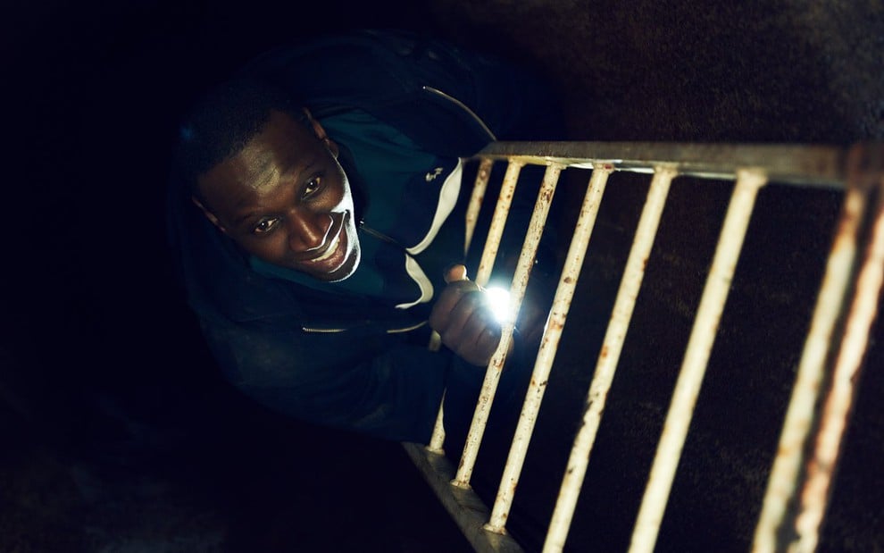 Assane Diop (Omar Sy) ri ao subir de escada em ambiente fechado e escuro segurando uma lanterna na mão direita