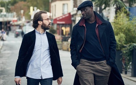 Benjamin Ferel (Antoine Goy) no lado esquerdo e Assane Diop (Omar Sy) no lado direito caminham pela rua e olham um para o outro em conversa