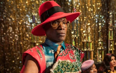 Pray Tell (Billy Porter) com olhar sério de óculos quadrado, chapéu vermelho e roupa com diversos adornos com fundo dourado desfocado