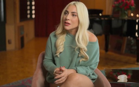Lady Gaga sentada em poltrona, com as pernas cruzadas, cabelo loiro sobre os ombros e blusa verde