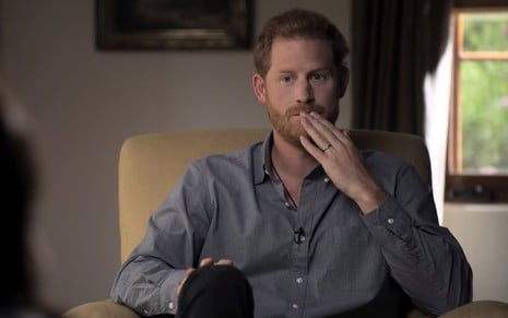 Príncipe Harry de camisa cinza sentado em poltrona com a mão esquerda na altura da boa com expressão séria