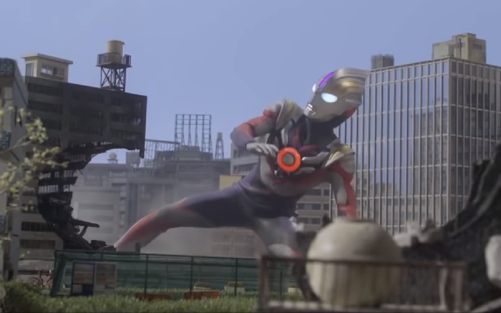 Ultraman Orb com roupa vermelha com armadura prata em posição de defesa quase deitado entre os prédios destruídos