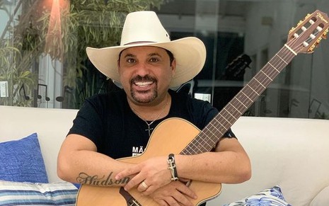 Cantor Edson de camiseta preta, calça jeans, chapéu branco, segurando um violão