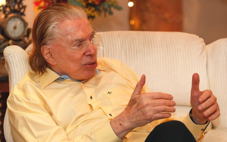 De cabelo branco e sentado em uma cadeira, Silvio Santos conversa em casa