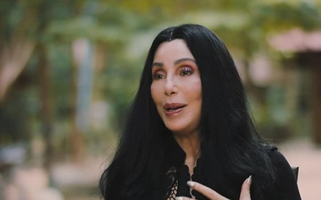 Cher está com longos cabelos pretos, e toda de preto, dando entrevista em um cativeiro de um elefante em cena de Cher e o Elefante Solitário
