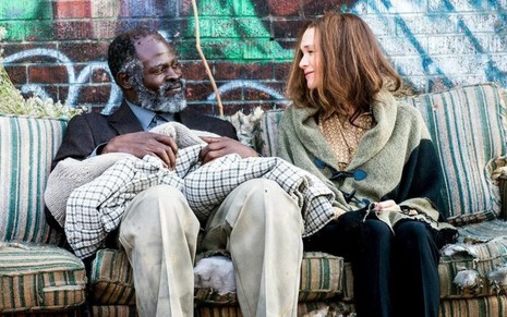 Sentados em um sofá rasgado, Djimon Hounsou e Renée Zellweger se olham sorrindo em cena de Somos Todos Iguais (2017)